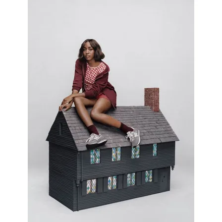 A New Balance ad creative: Ayo Edebiri sitting on the roof of a dark model house in a burgundy tracksuit and grey 574 trainers