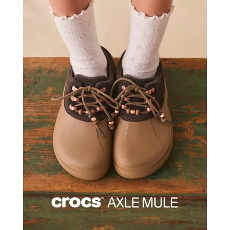 A Crocs ad creative: a pair of tan and brown Axle Mules worn with frilled white socks on a weathered green and brown wooden floor