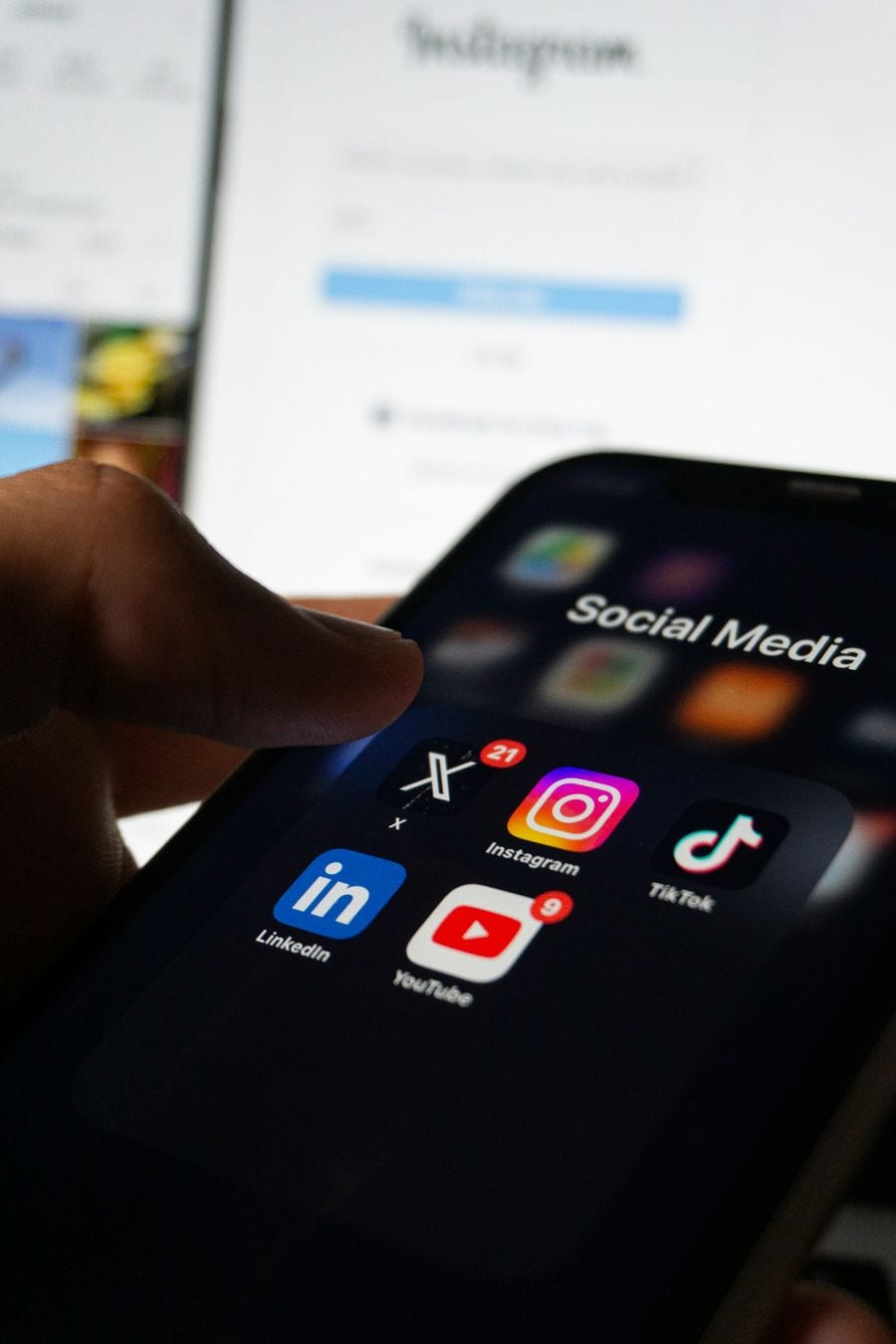 A hand holds a phone with a Social Media folder open, showing LinkedIn, Instagram, YouTube and TikTok icons, in front of a laptop