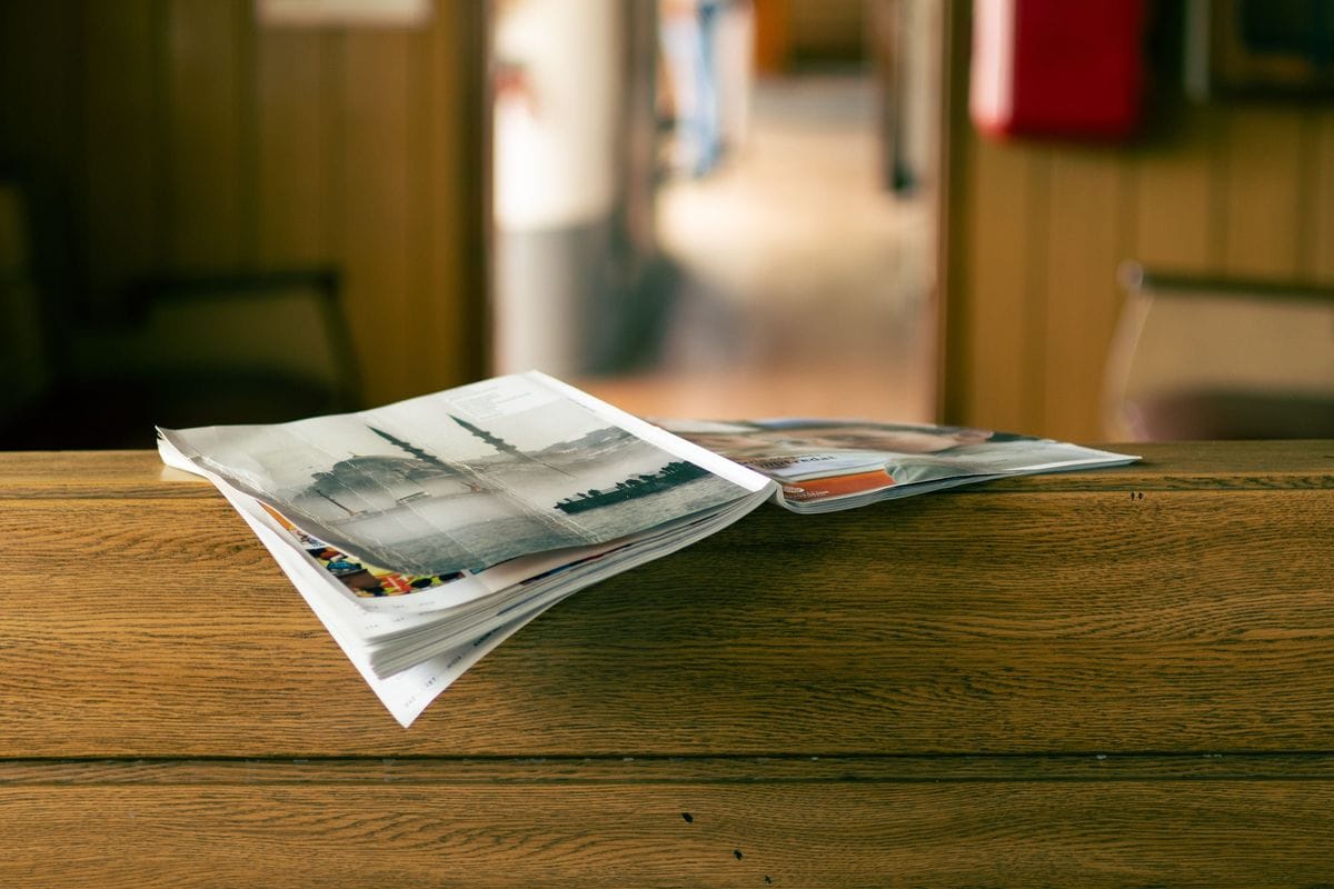 Open magazines lie on a wooden table in a quiet cafe: in print, the advertisement feature label sits above the headline, and the same rule holds on a phone