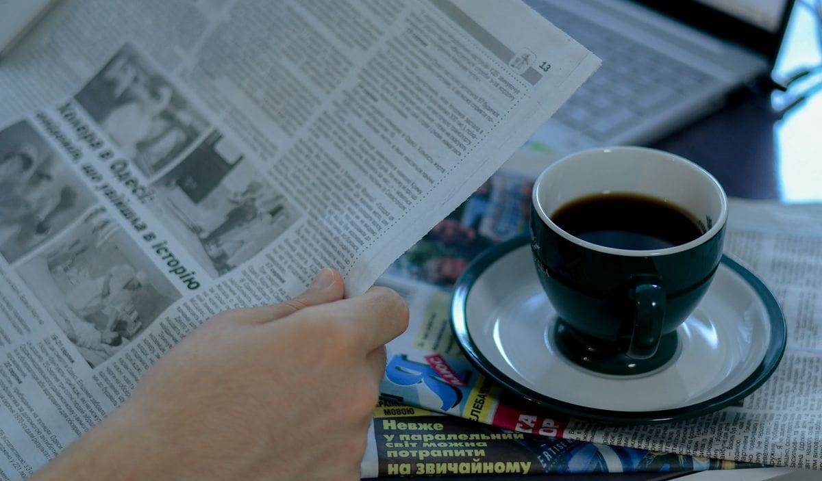 A reader holds an open newspaper next to a cup of black coffee: the editorial setting every advertorial borrows its look from