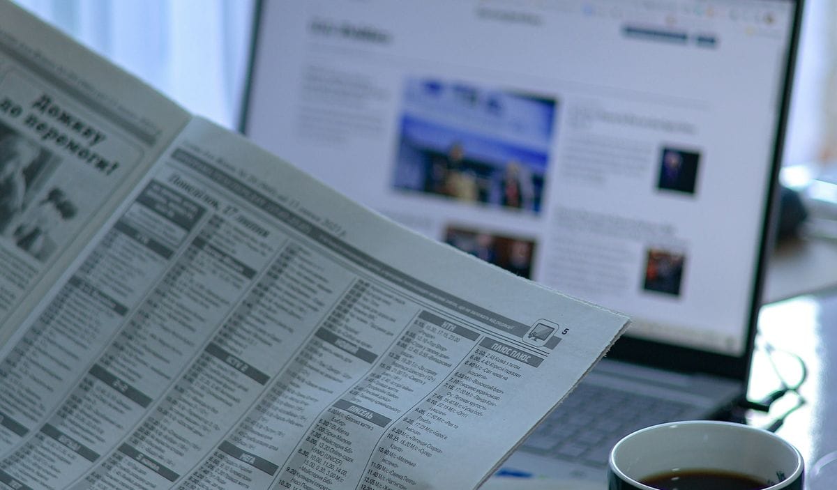 A newspaper held in front of an open laptop and a coffee: the advertorial borrows the look of print, never a real newspaper's name