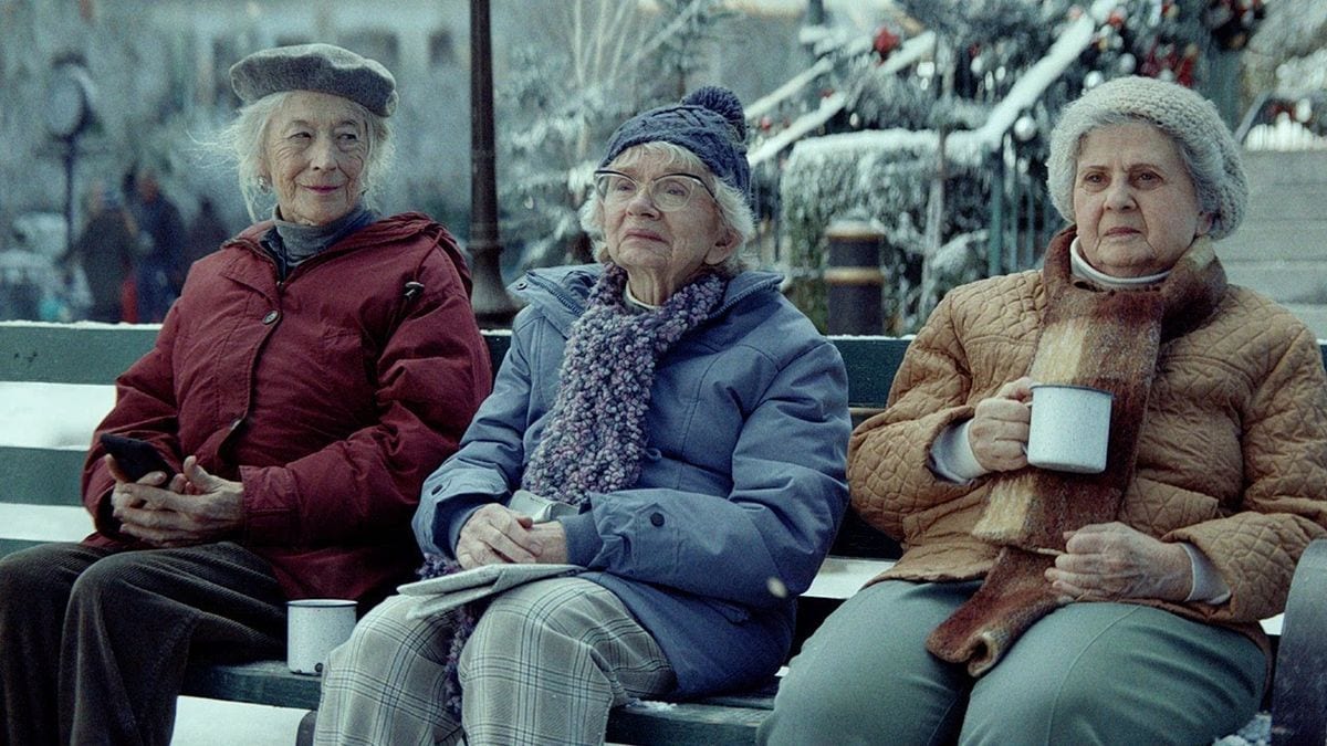 Amazon's 2023 Christmas ad Joy Ride: three older women in winter coats sit on a snowy park bench, holding tin mugs, watching others sled