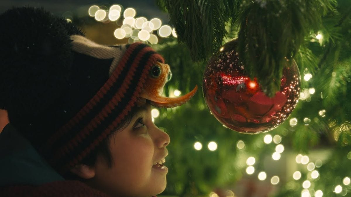 Disney's 2024 holiday short The Boy & The Octopus: a boy with a small orange octopus on his knitted hat looks up at a red bauble on a lit Christmas tree