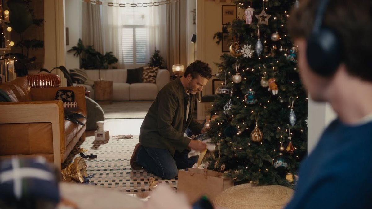 John Lewis's 2025 Christmas ad Where Love Lives: a dad kneels by the Christmas tree among torn wrapping paper while his son watches from the sofa