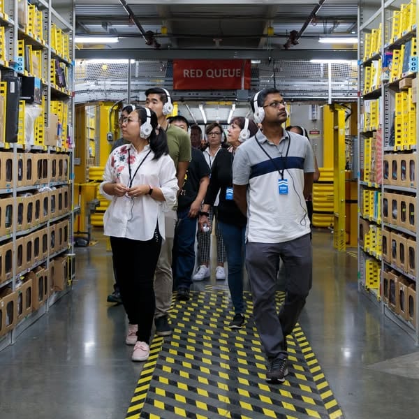 An ad by Amazon FC Tours in Meta's Ad Library: visitors in headsets walk between yellow shelves in a fulfillment center, inviting people to go behind the scenes on a free tour