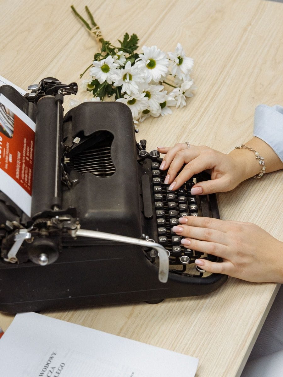 Hands type on a vintage typewriter next to white flowers: catalog copy that sells a scene still has to be written one sentence at a time