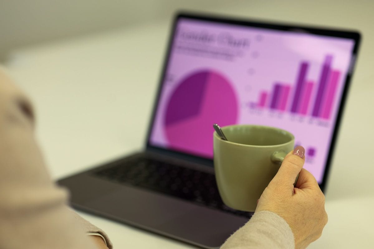 A hand holding a coffee cup next to a laptop showing sales charts: Meta never charges more than the budget you set, but the budget says nothing about the price of a click