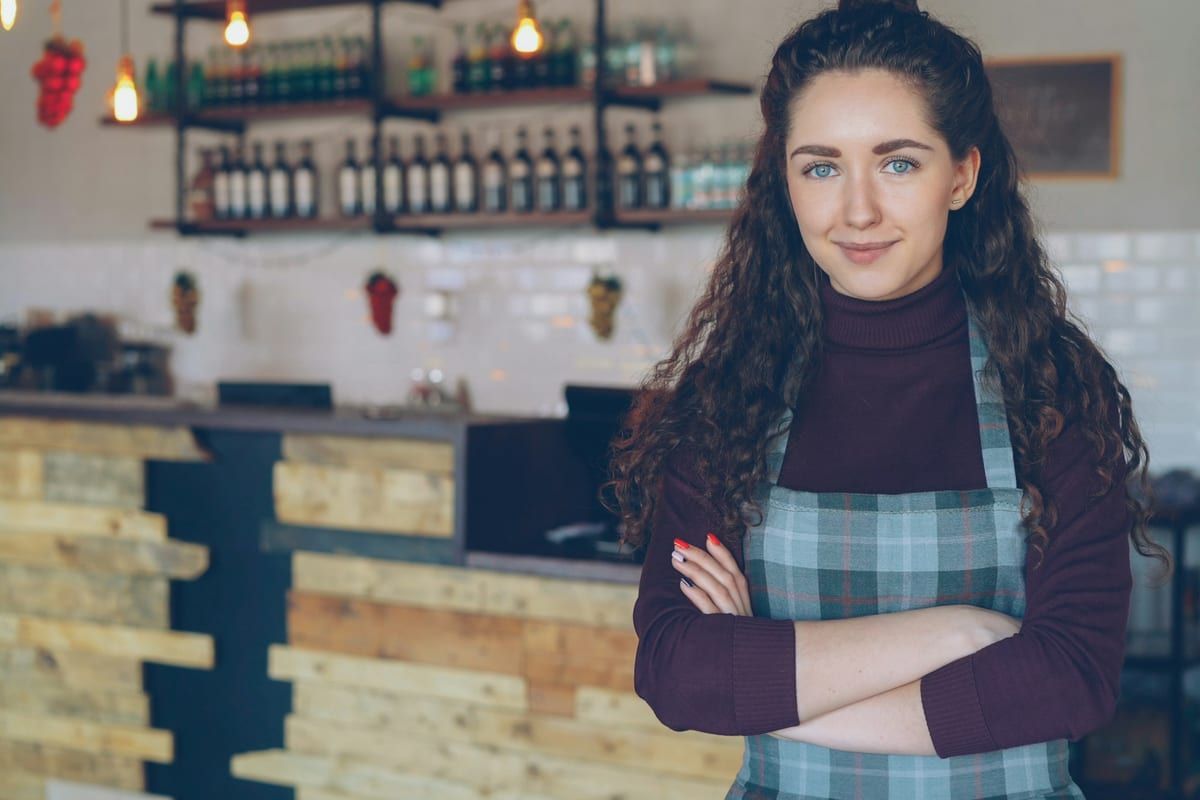 A smiling cafe owner in an apron standing with her arms crossed in front of her counter: most Facebook pages that work are run by the person who serves the customers