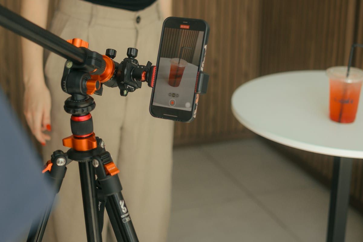 A phone on an orange tripod filming a woman behind a cafe counter: one person with a phone can film most Reel ideas in a few minutes