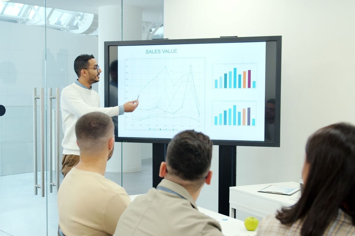 A man presenting a sales chart on a large screen to colleagues in a meeting room: blended revenue and ad spend side by side tell you whether the ads pay