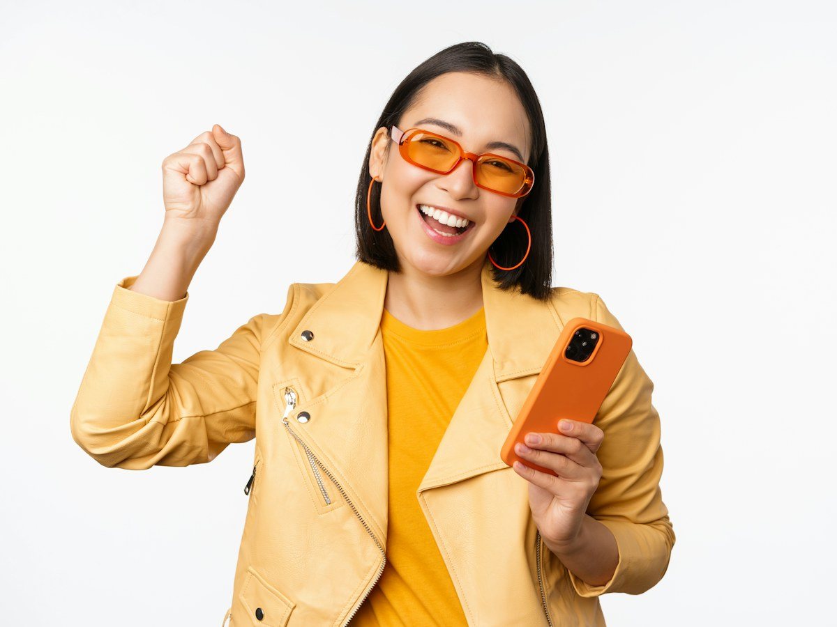 A woman in a yellow jacket celebrating at her phone: a caption that asks a real question is the one that brings replies