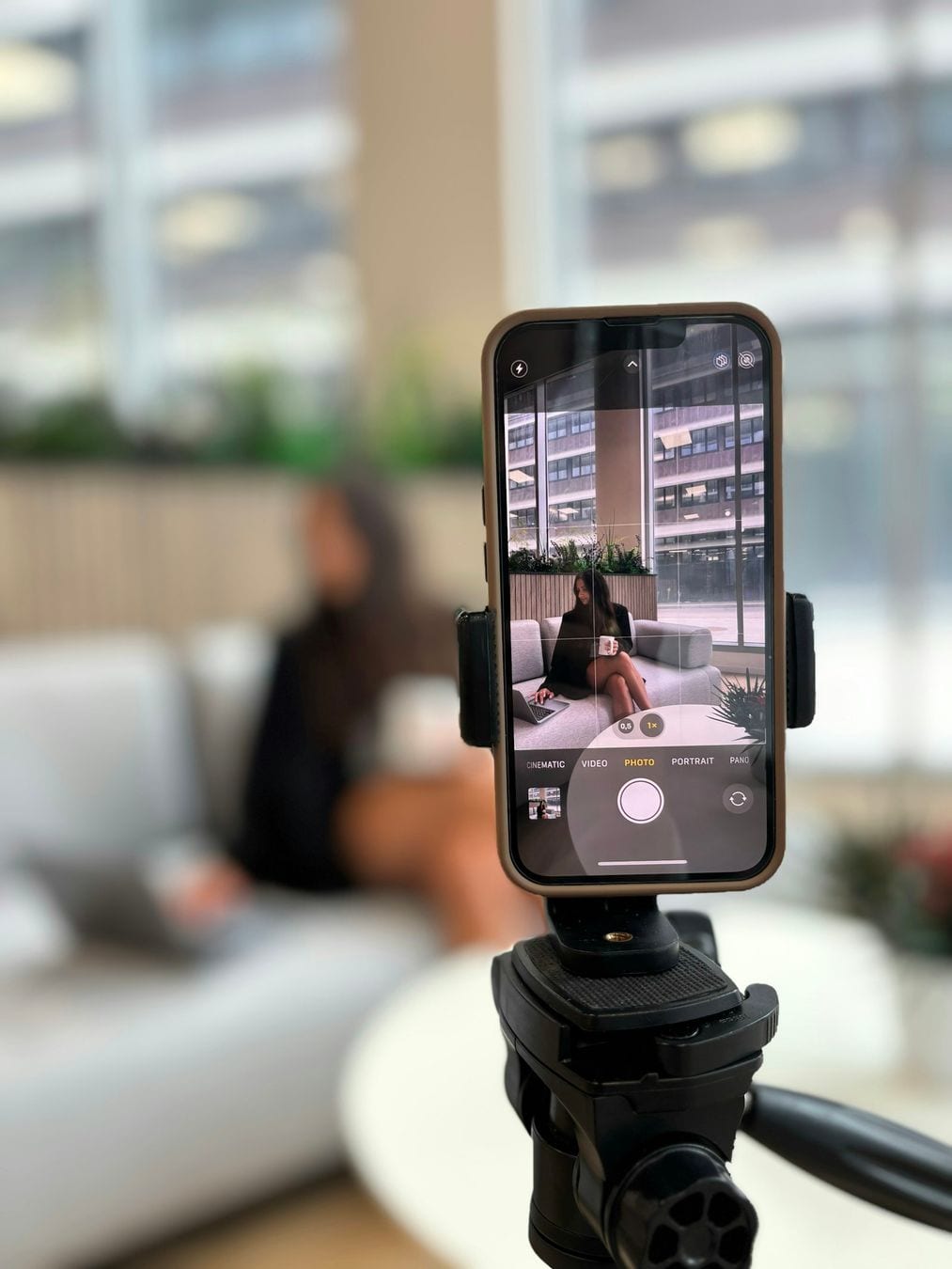 A phone on a tripod films a woman on a white sofa in a bright room, the set-up for a customer video testimonial