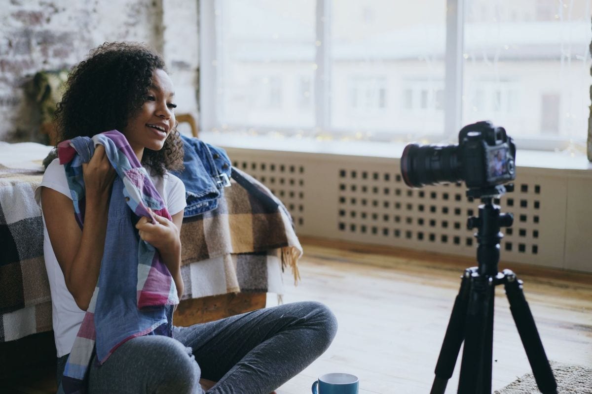 A young woman sits on her bedroom floor holding a scarf up to a camera on a tripod: the kind of at-home try-on a DTC fashion brand books a UGC creator for