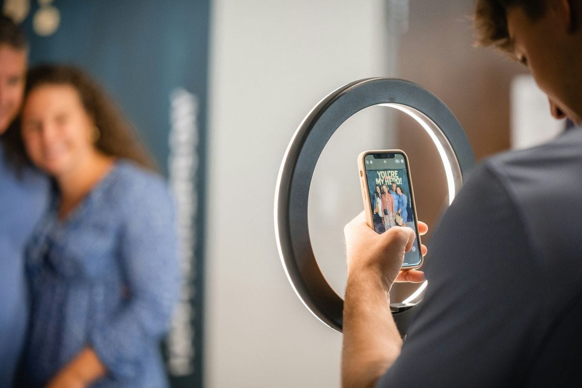 A man holds a phone in front of a ring light and frames two smiling people: footage shot by the creator, which a Spark Ad or partnership ad then runs under the creator's name