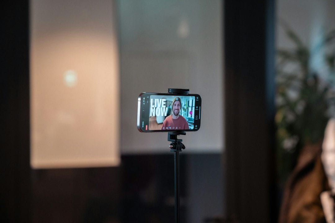 A phone on a tripod records a live video in a dim room, with a man on its screen