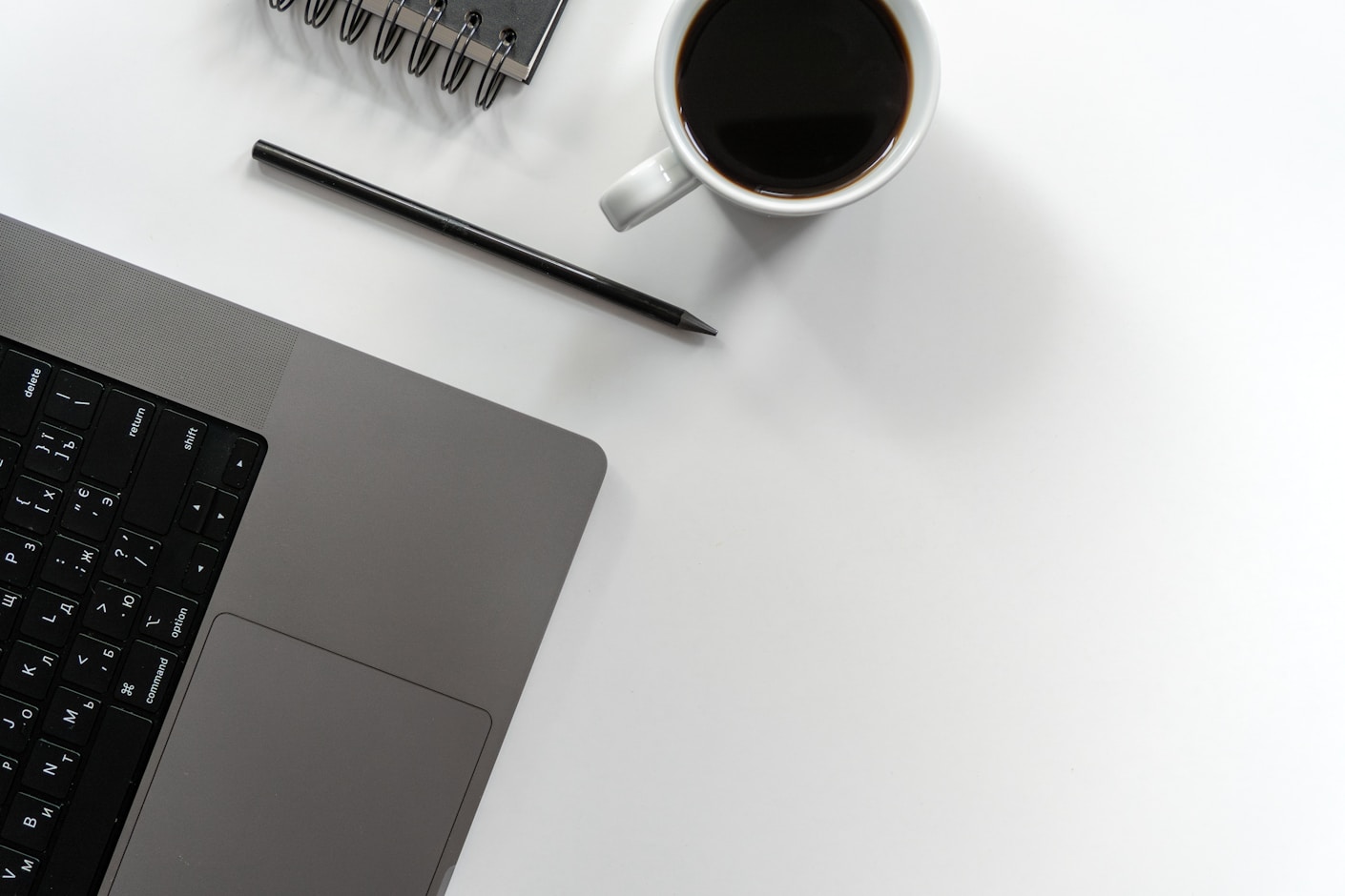 A closed laptop, a cup of coffee, a pen and a notebook on a plain white desk