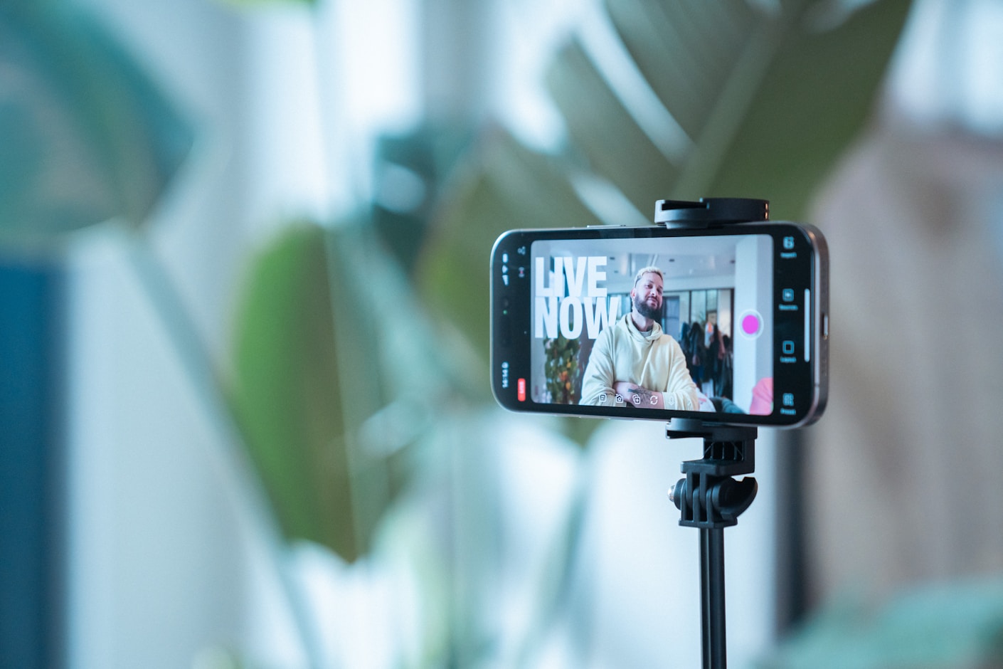 A phone on a tripod recording two people, the screen showing a LIVE NOW overlay, with plants behind it