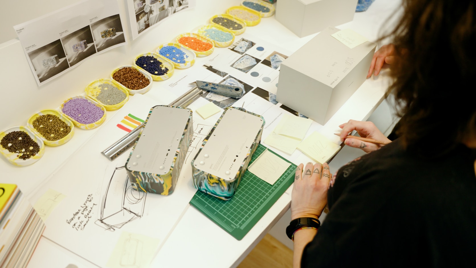 A designer at a studio table working through trays of colour samples and packaging models, with notes and a cutting mat