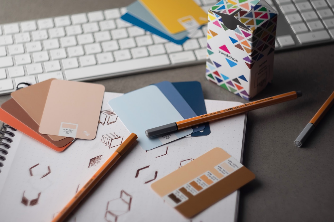 A designer desk with colour swatch cards in coral and blue, pencils, a patterned box and a keyboard