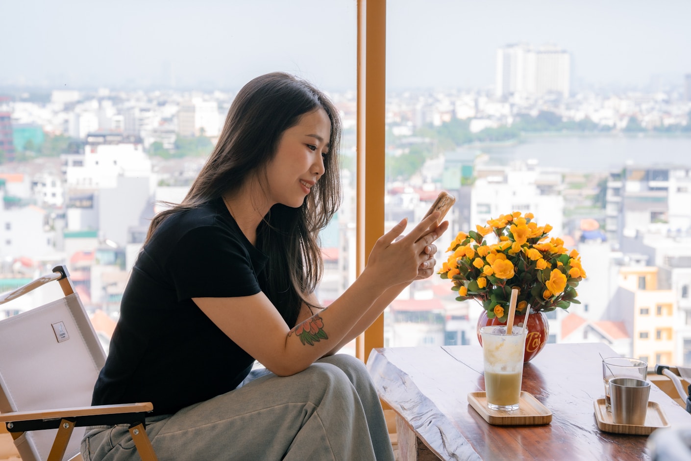 A woman sitting by a wide window with a city view, looking at her phone over an iced coffee