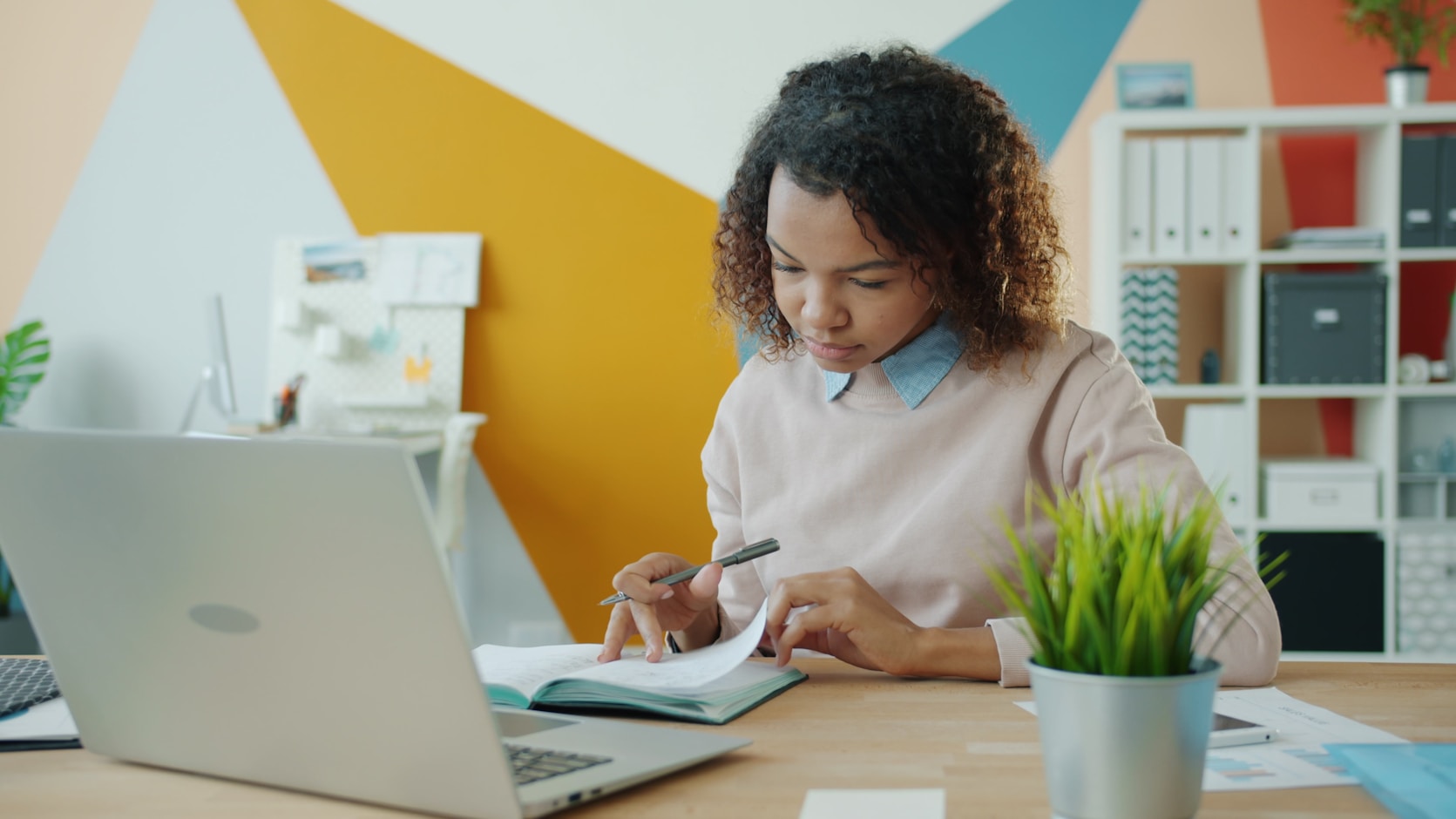 A woman at a desk writing in a notebook beside an open laptop, comparing what is on the screen against her notes