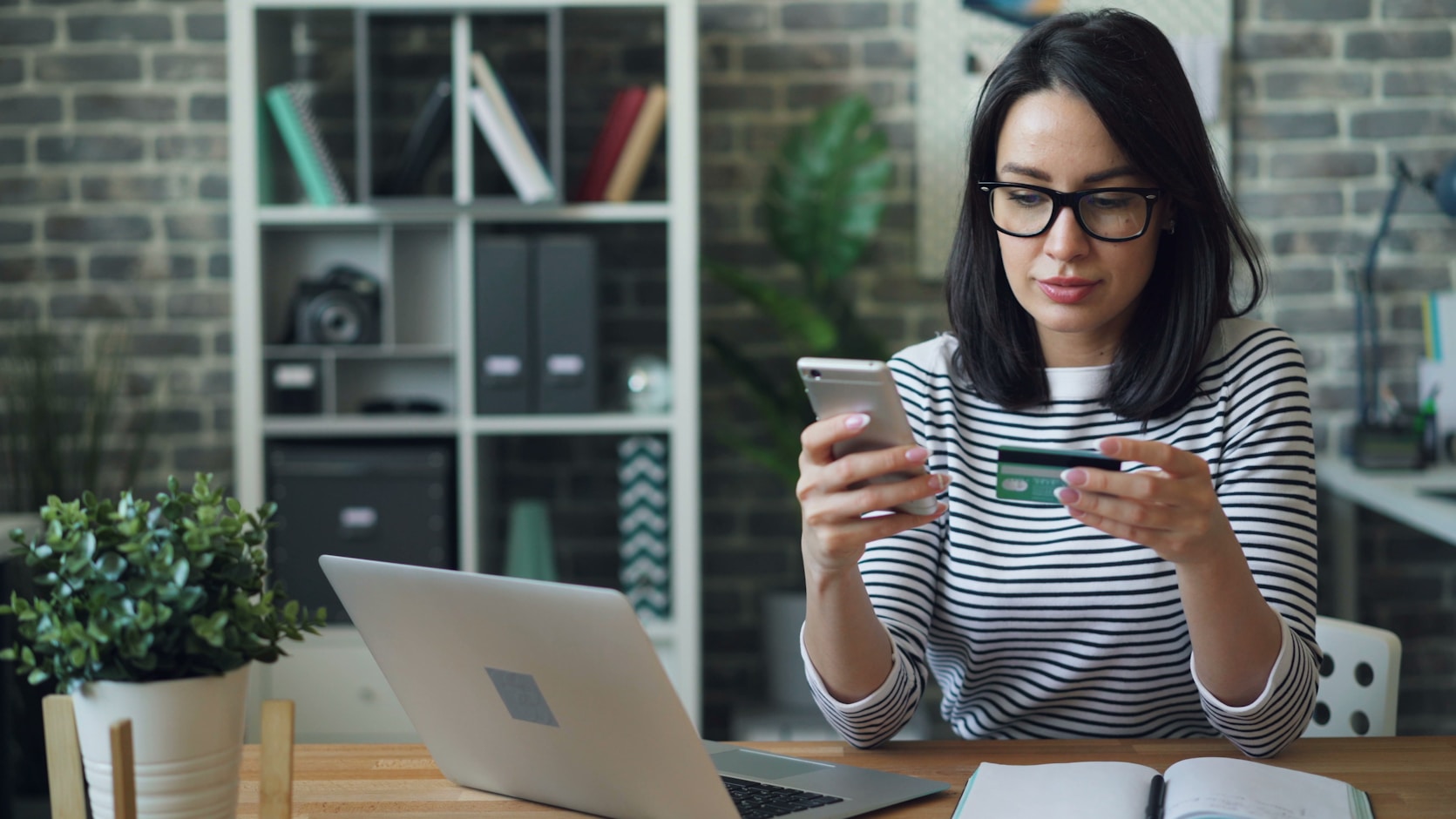 A woman holding a bank card in one hand and her phone in the other, in front of an open laptop