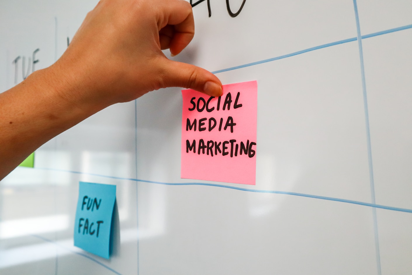 A hand placing a pink sticky note reading SOCIAL MEDIA MARKETING onto a whiteboard grid of days, beside a blue note reading FUN FACT
