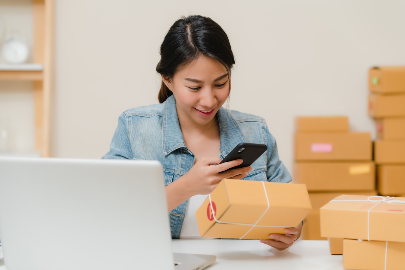 A woman checking a parcel against her phone at a desk stacked with packed boxes